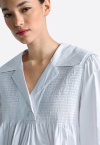 Fehér ing, gallérral, ráncolt törzzsel és pöndörített ujjakkal. Az anyag sima, enyhén fényes és laza szabású. Két gomb a nyakrésznél.