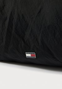 Fekete szövet, texturált felület, amely egy téglalap alakú logócímkét tartalmaz, rajta "TOMMY JEANS" felirat fehér, piros és kék színekben.