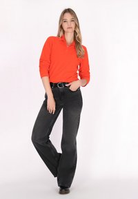 Oranje poloshirt met lange mouwen en een rits aan de voorkant, gecombineerd met hoge zwarte flared jeans. Glad materiaal, relaxte pasvorm, met een zwarte riem.