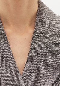 Šedý sako s herringbone vzorem a klopovým límcem, vyrobené z texturované látky s jemným leskem. Detailní záběr na výstřih ukazuje precizní prošívání.