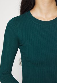 Tyrkysový pletený top s dlouhými rukávy a kulatým výstřihem, který má texturovaný vzor. Materiál vypadá měkký a dobře se přizpůsobuje postavě.