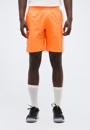 Άνδρας με φωτεινό πορτοκαλί αθλητικό σορτς, λευκές κάλτσες και μαύρα αθλητικά παπούτσια, στέκεται μπροστά σε απλό φόντο.