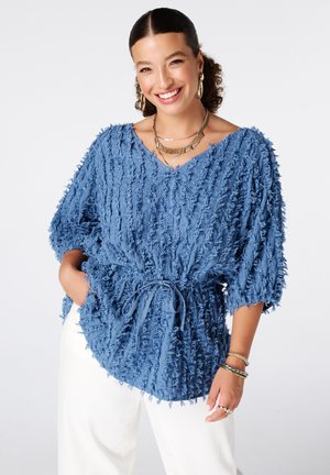 Vrouw glimlachend met een gestructureerde blauwe V-hals top met een tailleband en witte broek, geaccentueerd met gouden sieraden en armbanden.