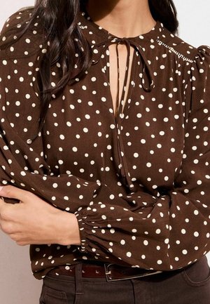 Frau trägt eine braune Bluse mit weißen Polka-Punkten, langen Puffärmeln und einem gebundenen Schlüsselloch-Ausschnitt, kombiniert mit dunkler Hose und Gürtel.