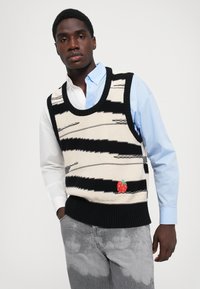 Un jeune homme portant un gilet en sweat noir et blanc à motifs avec un patch de fraise rouge, une chemise bleu clair et blanche, et un jeans gris délavé.
