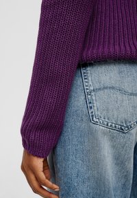 Fialový pletený svetr s žebrovanou texturou a přiléhavým lemem na rukávech. Nošený s světle modrými, poničenými džínami, které mají detail zadní kapsy.