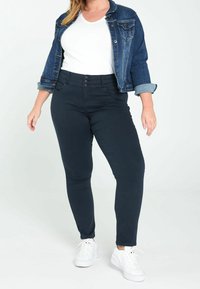 Jean skinny en denim foncé à taille haute, avec une braguette à boutons et quatre poches, porté avec un t-shirt blanc et une veste en denim.