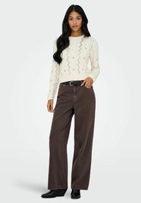 Pull en maille torsadée crème avec des accents floraux, associé à un pantalon marron ample et des bottes à talons compensés noires, présenté sur un fond minimal.