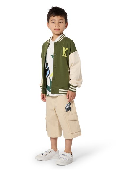Veste universitaire verte et crème avec la lettre "K", t-shirt graphique blanc, shorts cargo beige avec motif d'ours, et chaussures blanches à brides.