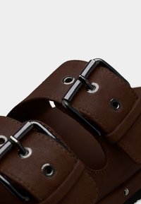 Καφέ δερμάτινα σανδάλια με δύο ρυθμιζόμενους ιμάντες, μεταλλικά ασημένια αγκράφια και μεταλλικές οπές πάνω σε μαλακή, υφή σόλα.