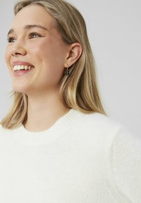 Pull en maille léger de couleur crème avec un col rond et des poignets côtelés. Présentant de petites boucles d'oreilles noires au design circulaire.