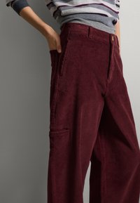Pantaloni di velluto a coste color bordeaux con texture a coste verticali, gamba larga e tasche laterali. Abbinati a un maglione a righe grigio.