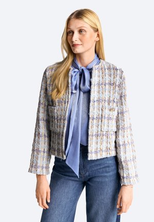 Blonde Frau trägt eine strukturiert gestreifte Jacke in Blau und Beige über einer seidenen blauen Bluse mit einer Schleife, kombiniert mit hoch taillierten blauen Jeans.