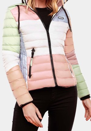 Kobieta w pikowanej kurtce z pastelowymi blokami kolorów, w tym różowym, zielonym, białym i niebieskim, zapinanej na suwak na czarną bluzkę, z rękami swobodnie opuszczonymi.