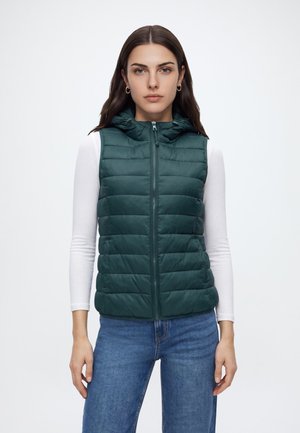Femme portant une veste sans manches matelassée vert foncé sur une chemise blanche à manches longues et un jean bleu, debout devant un fond uni.