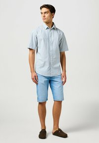 Camicia a maniche corte con bottoni a strisce blu e bianche, abbinata a pantaloni corti in denim blu chiaro e scarpe slip-on marrone scuro.