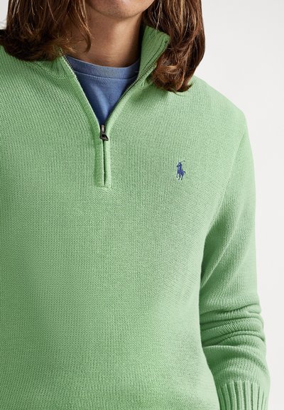 Világoszöld cipzáras pulóver texturált kötött mintával, a mellkasán egy kis kék logóval és bordázott mandzsettákkal. Alatta egy kék ing látható.