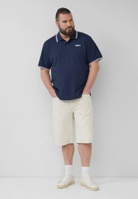 Dunkelblauses Poloshirt mit weißen Akzenten, kombiniert mit cremefarbenen Shorts. Weiße Sneakers und Socken, stehend mit Händen in den Taschen vor einem neutralen Hintergrund.