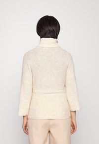 Pull en tricot de couleur crème avec un col montant, des manches trois-quarts et une ceinture nouée à la taille. Texture douce avec une coupe ample.
