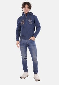 Felpa con zip color navy con cappuccio, caratterizzata da testi e disegni stampati in oro. Abbinata a jeans blu e sneaker beige.