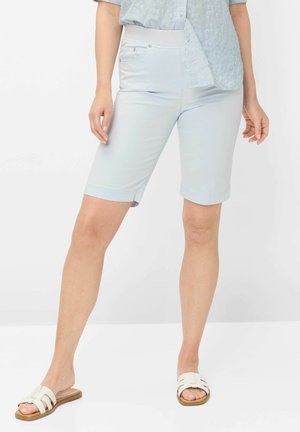 Frau trägt hellblaue knielange Shorts, hellblaues Hemd mit Knöpfen und weiße offene Sandalen und steht auf weißem Boden.