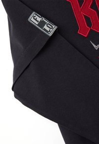 Primer plano de una tela negra con una letra roja bordada y una pequeña etiqueta rectangular con un diseño gráfico en blanco y negro.