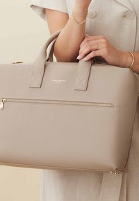 Lichtbeige leren handtas met gouden hardware en een voorzak met rits. Beschikt over korte handgrepen en een gestructureerd oppervlak. Merklogo zichtbaar.