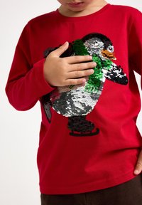 Red long-sleeve shirt featuring a textured penguin design made of colored sequins, differing shades of green, black, and white.