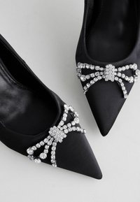 Melnas satīna augstpapēžu kurpes ar smailo purngalu un dekoratīvu rhinestone loku akcentu priekšpusē. Gluda tekstūra un izsmalcināts dizains.
