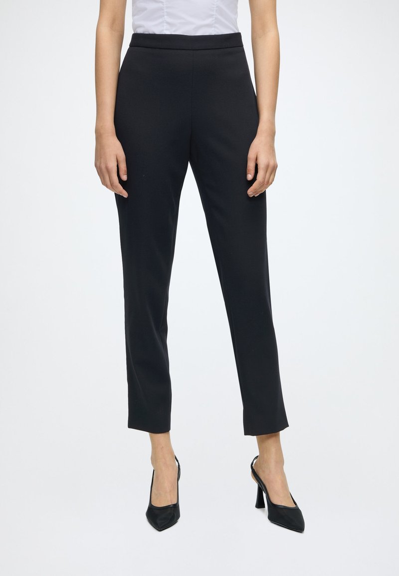 Femme portant un pantalon noir slim-fit chevilles, associé à des chaussures slingback noires à talons hauts et une chemise blanche rentrée.