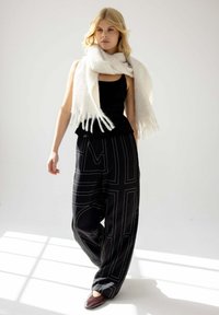 Pantalons larges noirs avec des motifs géométriques blancs, assortis à un haut noir ajusté et une écharpe blanche douce. L'ensemble est complété par des chaussures foncées.