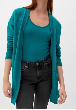 Otwarty kardigan w kolorze morskim noszony na morskim topie z dekoltem w łódkę, zestawiony z wysokim stanem czarnych jeansów na stojącej osobie.