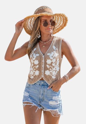 Gilet beige en maille avec broderie florale blanche, col en V, boutons sur le devant, porté sur des shorts en denim. Le modèle porte un chapeau de paille et des lunettes de soleil rondes.