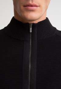 Suéter negro con cremallera y textura acanalada, con un cuello alto. Presenta una cremallera plateada en el frente para el cierre.