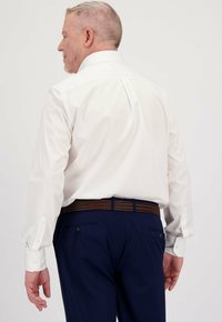 Man met kort haar in een witte blouse die in navyblauwe broeken is gestoken met een leren bruine riem, gezien van achteren.