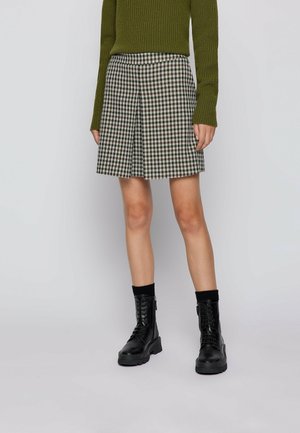 Femme portant une jupe pied-de-poule noir et blanc, un pull en maille vert olive, des chaussettes noires et des bottines noires à lacets.