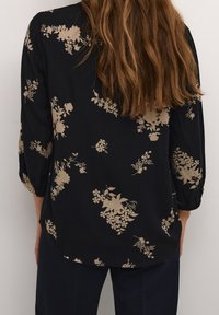 Blusa negra con estampado floral en tonos beige, con dobladillo redondeado y mangas arremangadas. Está hecha de una tela ligera con una textura suave.