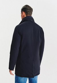 Czarny, średniej długości płaszcz z wysokim kołnierzem, o gładkiej fakturze i prostym kroju. Charakteryzuje się minimalnym przeszyciem i brakiem widocznych akcentów metalowych.