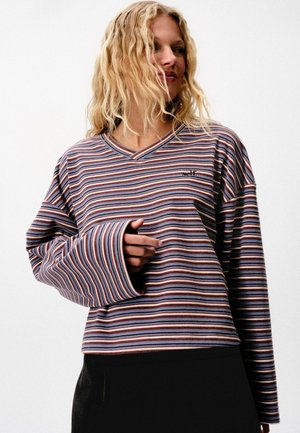 Blonde Frau mit welligem Haar trägt ein lockeres, langärmeliges gestreiftes Hemd mit der Aufschrift „self.“ auf der Brust, schwarze Hose und schaut seitlich.