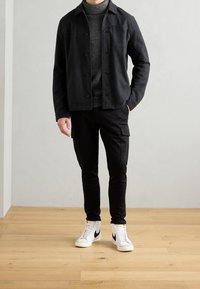 Černá košilová bunda z vlněné směsi přes šedý texturovaný rolák, doplněná černými cargo kalhotami a bílými vysokými teniskami s černými detaily.