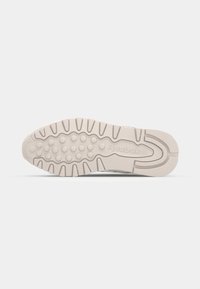 Reebok sneaker outsole in light beige, featuring a textured grip pattern with circular and linear elements. Durable rubber construction.