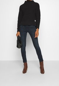 Suéter negro de punto con cuello alto, jeans ajustados azul oscuro, botines marrones y un bolso de cuero negro. Look sencillo y estilizado.