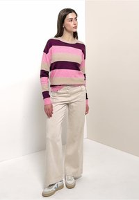 Frau in weit geschnittenen beigen Hosen, weißen Sneakers und einem langärmeligen gestreiften Pullover in Dunkelpurpur, Pink und Beige, die in einer Ecke steht.