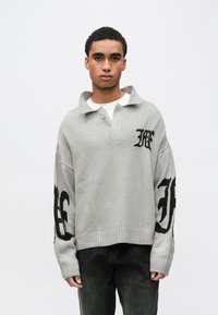 Γκρι πλεκτό πουλόβερ με γιακά και κουμπί, με μεγάλα μαύρα λογότυπα στα μανίκια. Το υλικό φαίνεται μαλακό και με υφή.