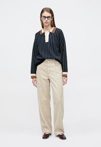 Γυναίκα με γυαλιά, μπλε ναυτικό μακρυμάνικο πόλο με ρίγες, μπεζ παντελόνι φαρδύ και σκούρα καφέ παπούτσια, που στέκεται μπροστά σε λευκό φόντο.