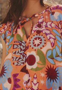 Blouse colorée à manches courtes en orange avec des motifs floraux et abstraits. Présente un décolleté en trou de serrure et un détail de nœud au col.