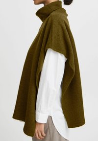 Poncho di lana verde oliva con colletto alto, vestibilità ampia e texture liscia, indossato sopra una camicia bianca con bottoni e pantaloni a righe.