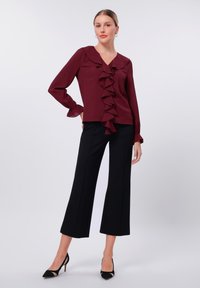 Burgundy blouse with ruffle detailing down the front, V-neckline, and long sleeves paired with black wide-legged trousers and black heels.