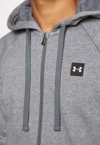 Felpa grigia con zip e tessuto texturizzato, dotata di cappuccio, cordini regolabili e un piccolo logo nero di Under Armour sul petto.