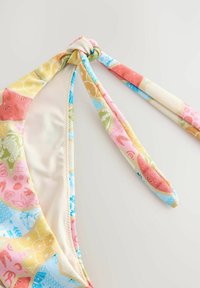 Daugiaspalvė bikinio viršutinė dalis su mazgo užrišimu. Raštai apima geometrines formas ir vaisius pastelinių atspalvių. Lygi audinio tekstūra.